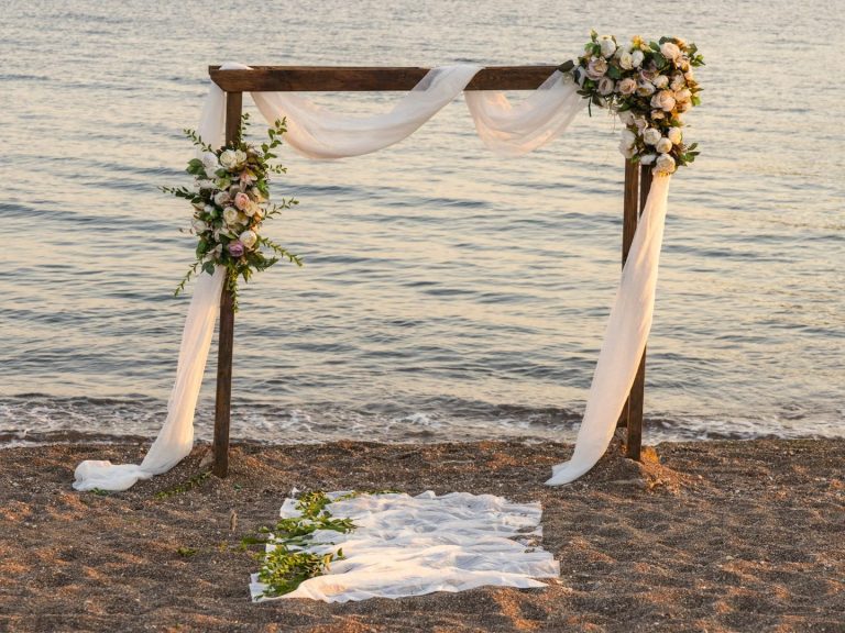 Hölzerner Hochzeitsbogen mit Blumen, weißem Tuch, Sandstrand und Meer.